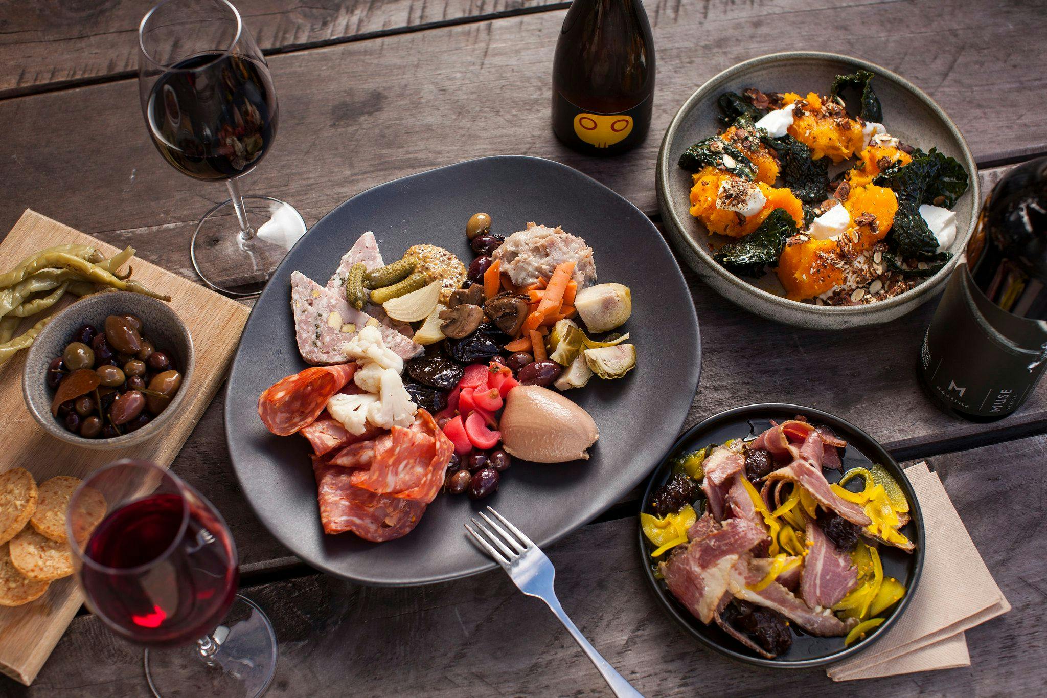 A wooden table with wine, Moo Brew, a ploughman's platter and dishes of pumpkin, charcuterie and olives