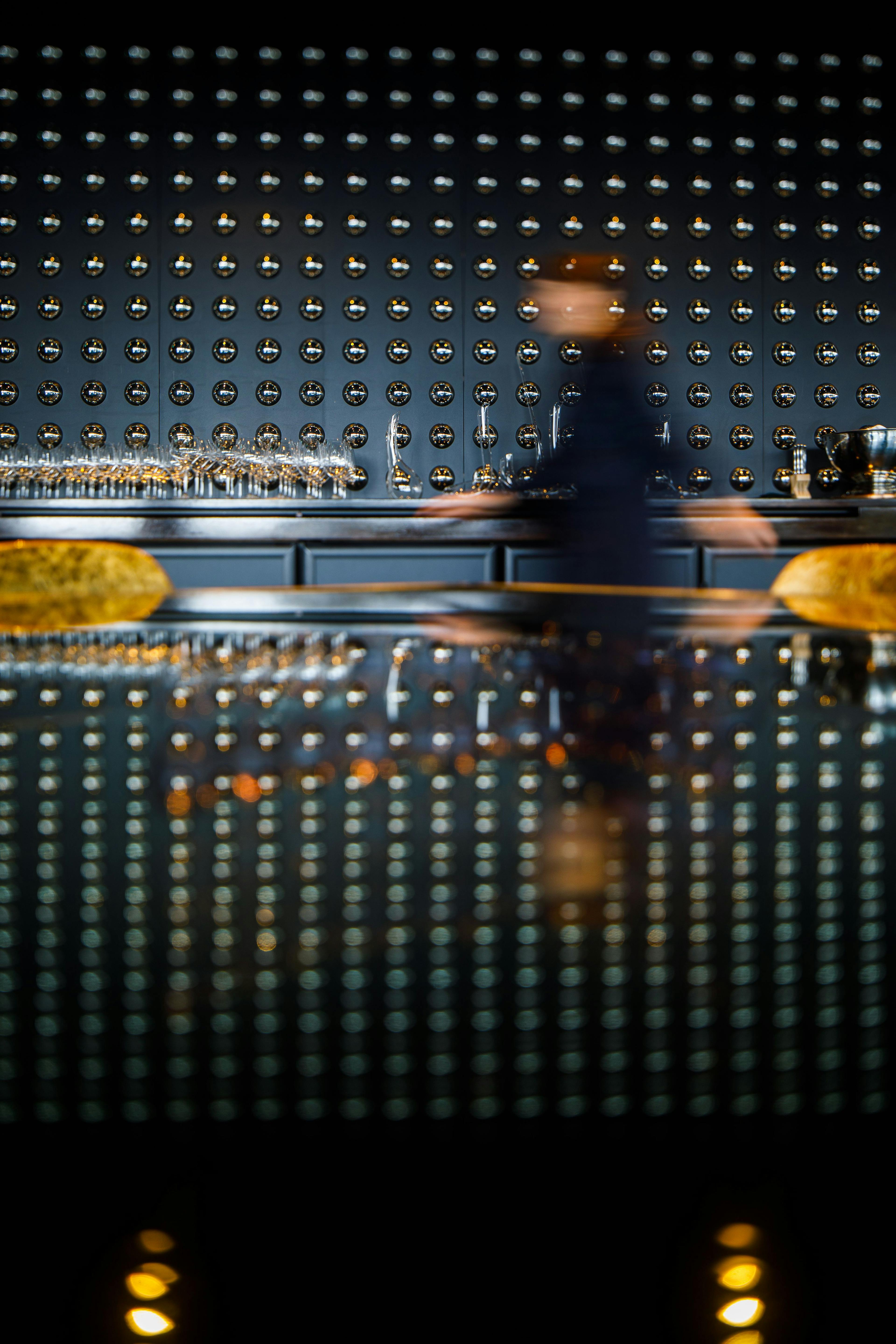 A waiter moves across a dark bar lined with dozens of wine glasses.