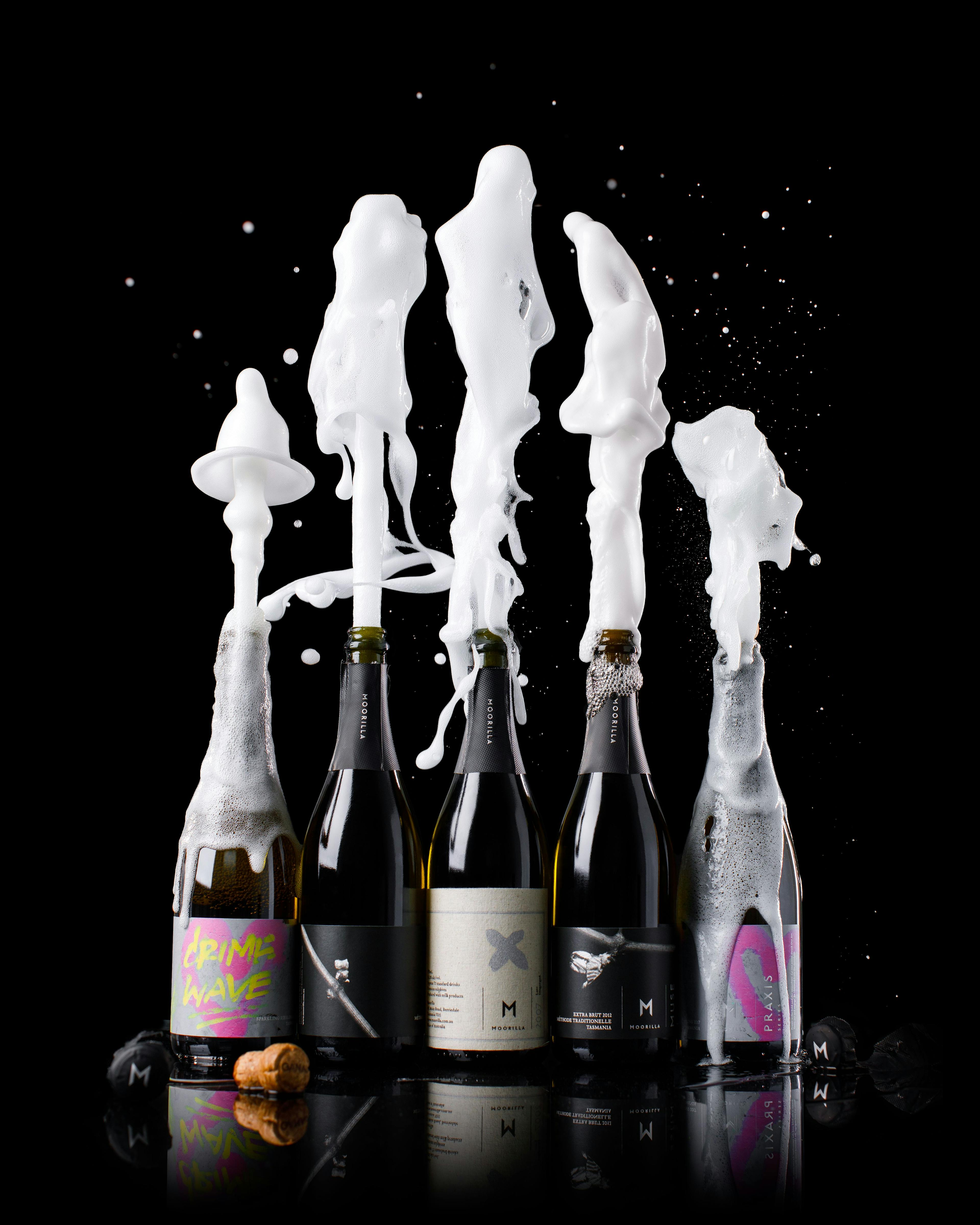 5 bottles of sparkling wine sit on a black background shooting plumes of fizzy wine