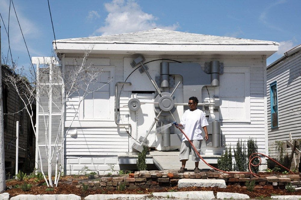 A person waters their garden in front of a white weatherboard house with what appears to be a vault lock as the entrance