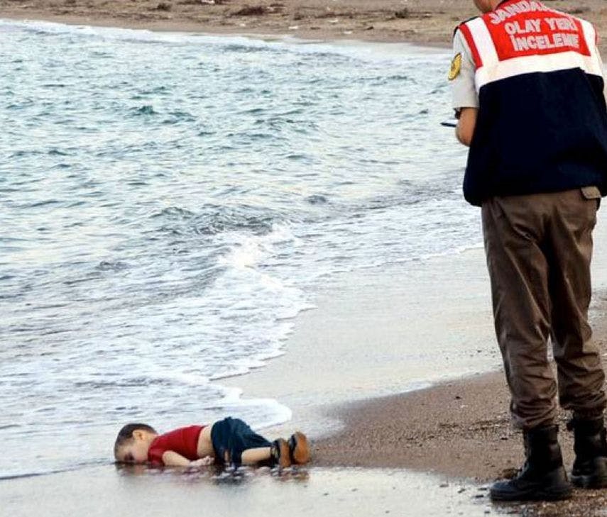 The deceased body of a small child lays face down on a beach. An officer stands above him writing in a notepad.